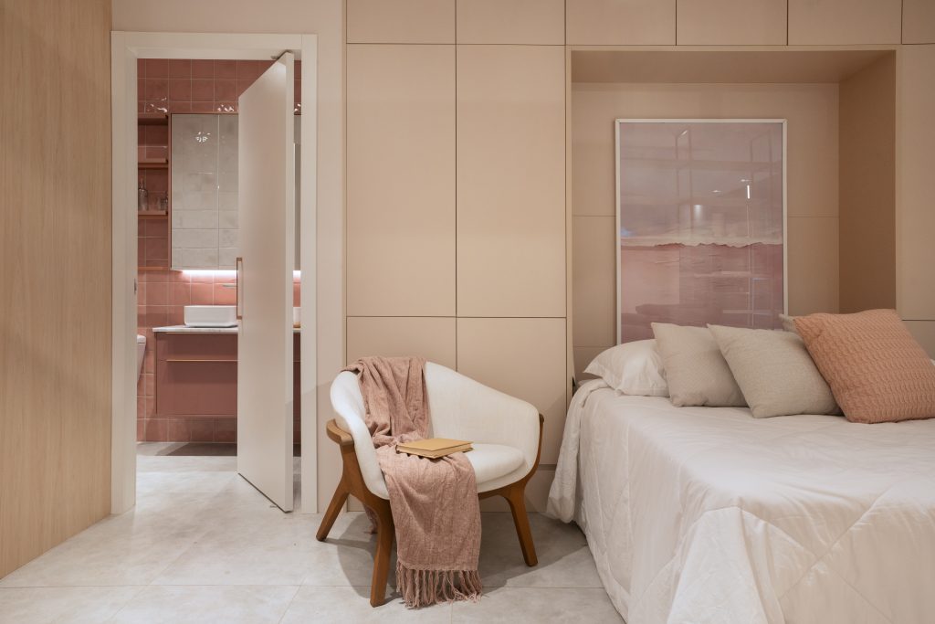 Quarto aconchegante em tons neutros com poltrona, manta, cama de casal e banheiro ao fundo.