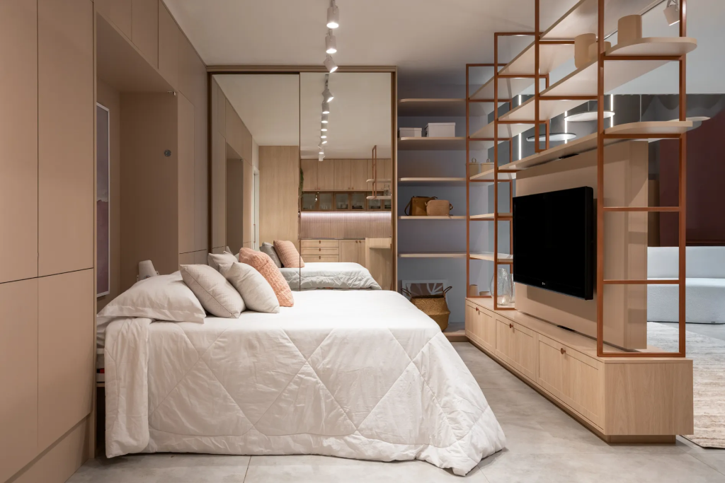 Quarto integrado a cozinha com cama de casal, armários espelhados e estante vazada com TV.