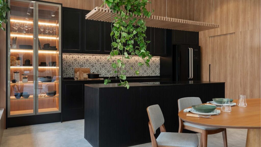Cozinha compacta com armários pretos, parede de madeira, ilha central com planta pendente, armário iluminado com louças expostas e mesa de jantar redonda com dois lugares posta para refeição.