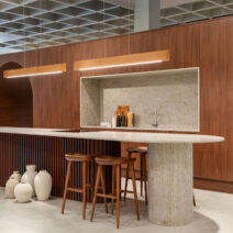 Cozinha com design sofisticado em madeira escura, bancada curva em pedra clara com banquetas, iluminação embutida e elementos decorativos no chão.