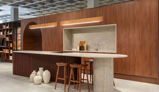 Cozinha com design sofisticado em madeira escura, bancada curva em pedra clara com banquetas, iluminação embutida e elementos decorativos no chão.