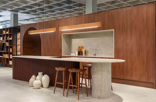 Cozinha com design sofisticado em madeira escura, bancada curva em pedra clara com banquetas, iluminação embutida e elementos decorativos no chão.